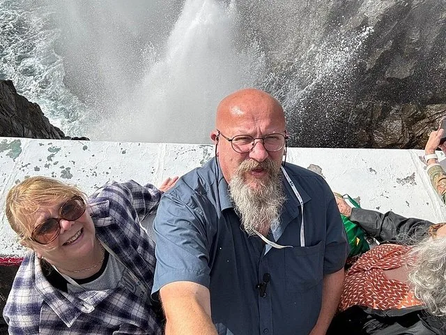 Ein Paar macht ein Selfie neben einem Blasloch, das Wasser in die Höhe spritzt.